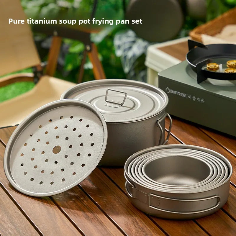 A titanium soup pot with lid, steamer tray, and a set of nested frying pans on a table.