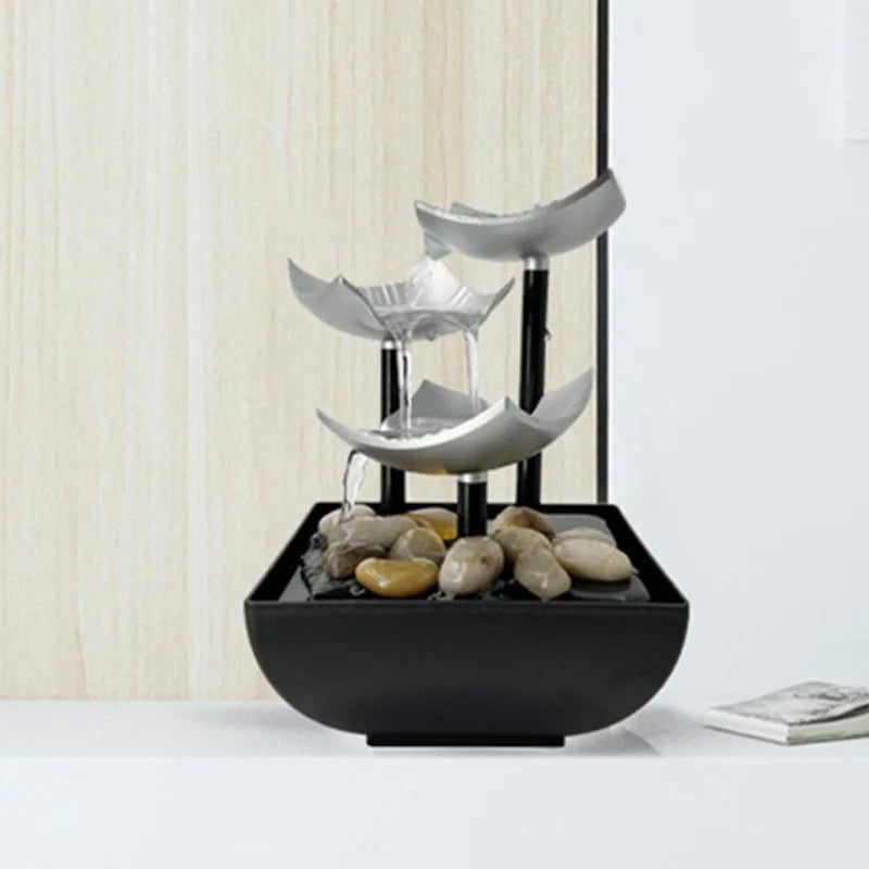 Modern three-tier tabletop water fountain with silver lotus-shaped bowls, black supports, and smooth decorative stones.