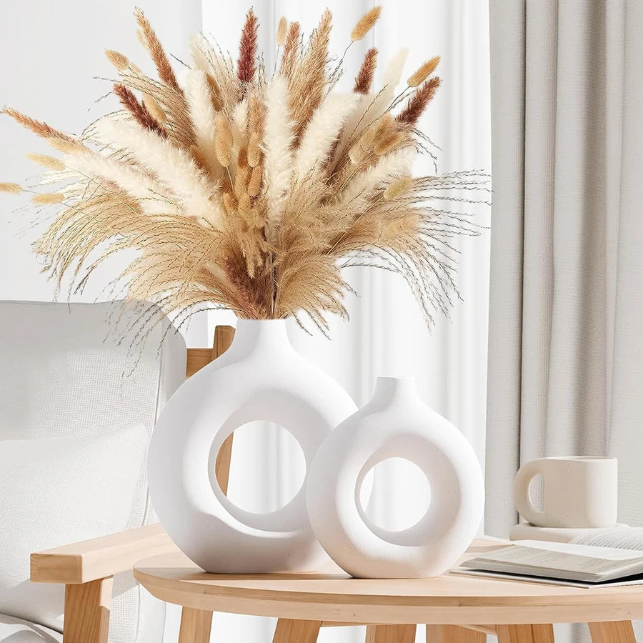 Two modern, white circular doughnut-shaped vases on a wooden table, featuring a large bouquet of dried pampas grass.