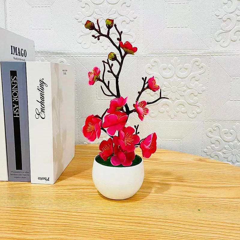Artificial pink plum blossoms in a small white pot, sitting on a wooden surface next to two white books.