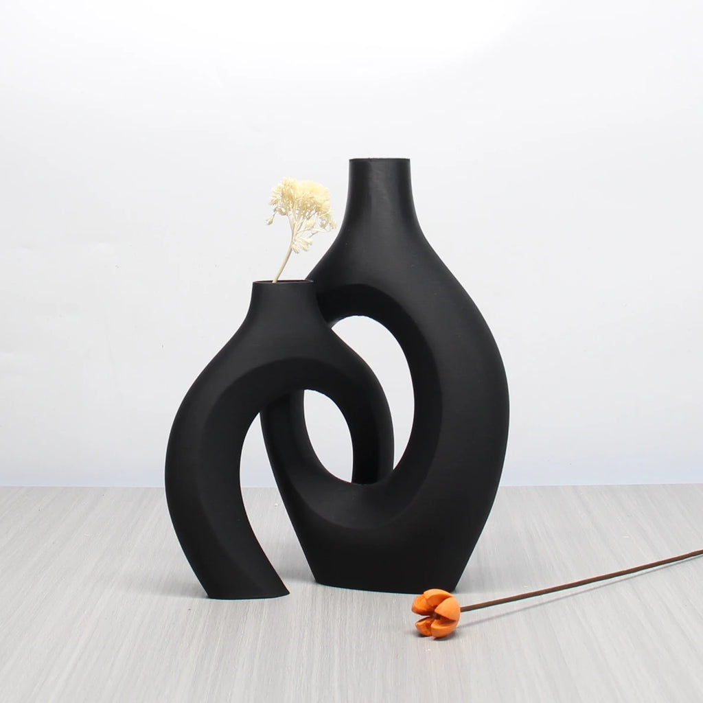 Two matte black, sculptural boho-style vases with flowing, curved silhouettes and cut-out centres on a wooden surface.