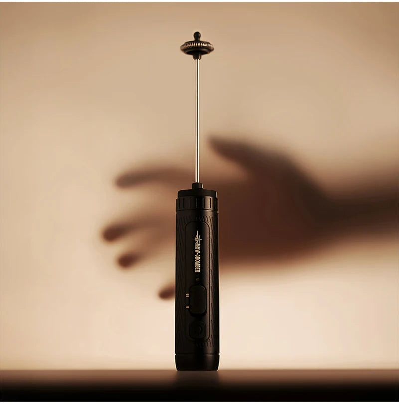 Handheld electric milk frother in matte black with a slim metal wand and whisk, set against a soft, lit background.
