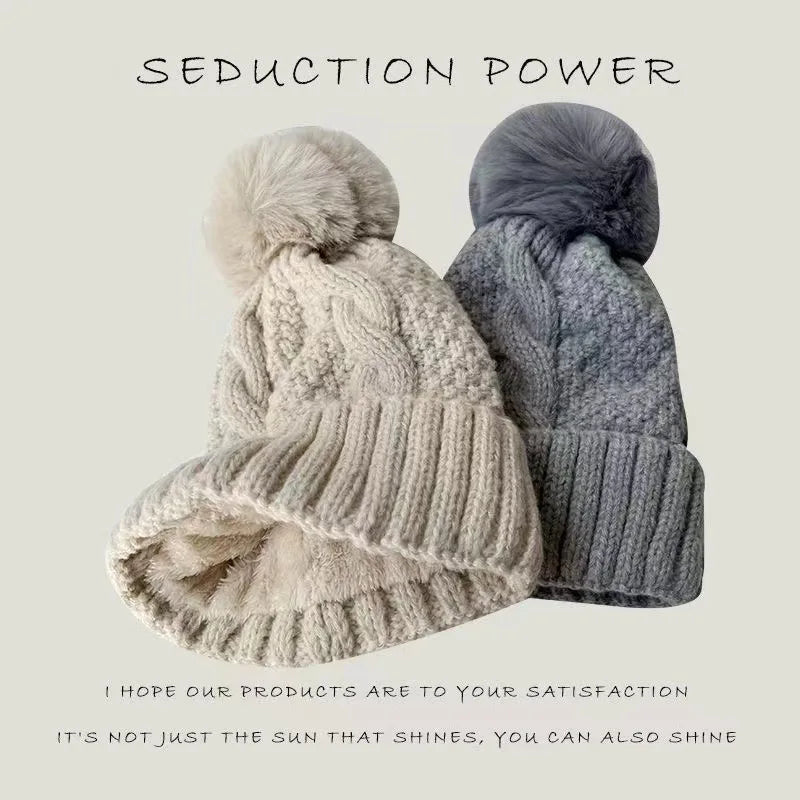 Two cable-knit winter beanie hats with faux fur pom-poms, one in beige and one in grey, with ribbed turn-up hems.