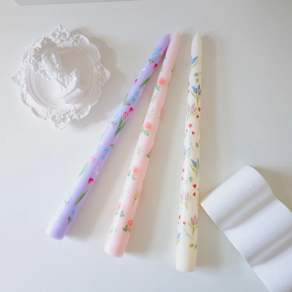 Three tall, tapered dinner candles in lilac, pink, and cream, featuring delicate floral prints and botanical designs.