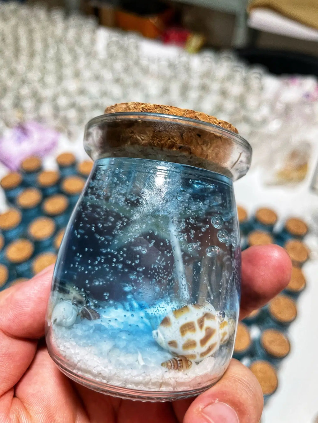 Small glass jar with a cork lid, featuring blue gel, white sand, and decorative seashells for a beach-themed display.