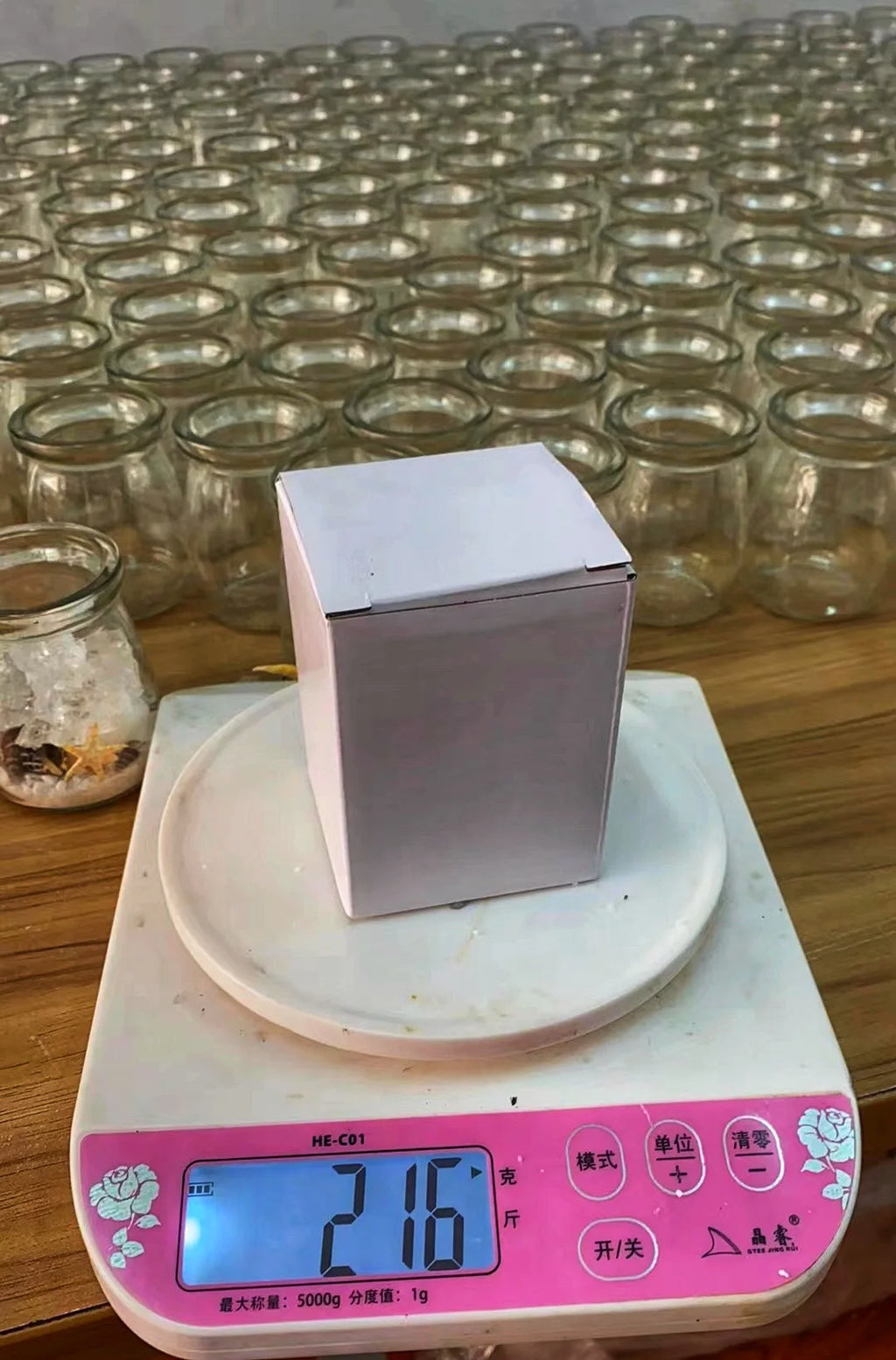 A plain white square box sitting on a digital kitchen scale, surrounded by many empty clear glass jars.