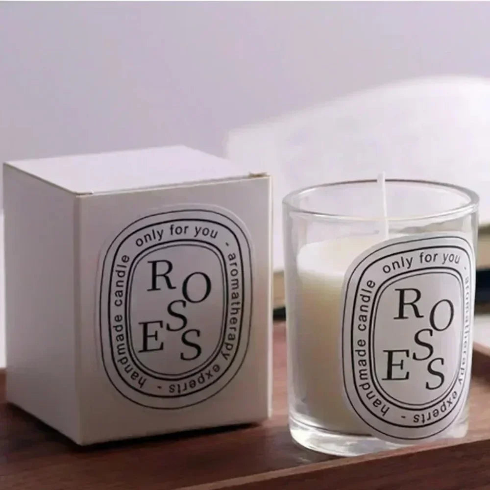 A white scented candle in a glass votive with a matching white box, both featuring minimalist "Roses" label designs.