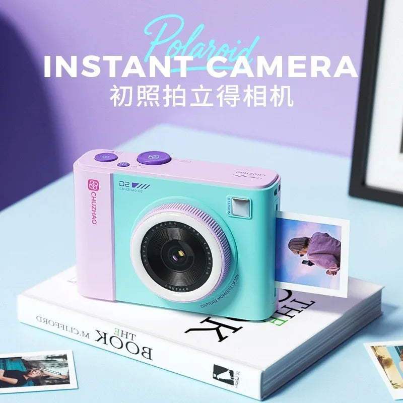 Pastel teal and purple Chuzhao D2 instant camera sitting on a white book, ejecting a printed photo.