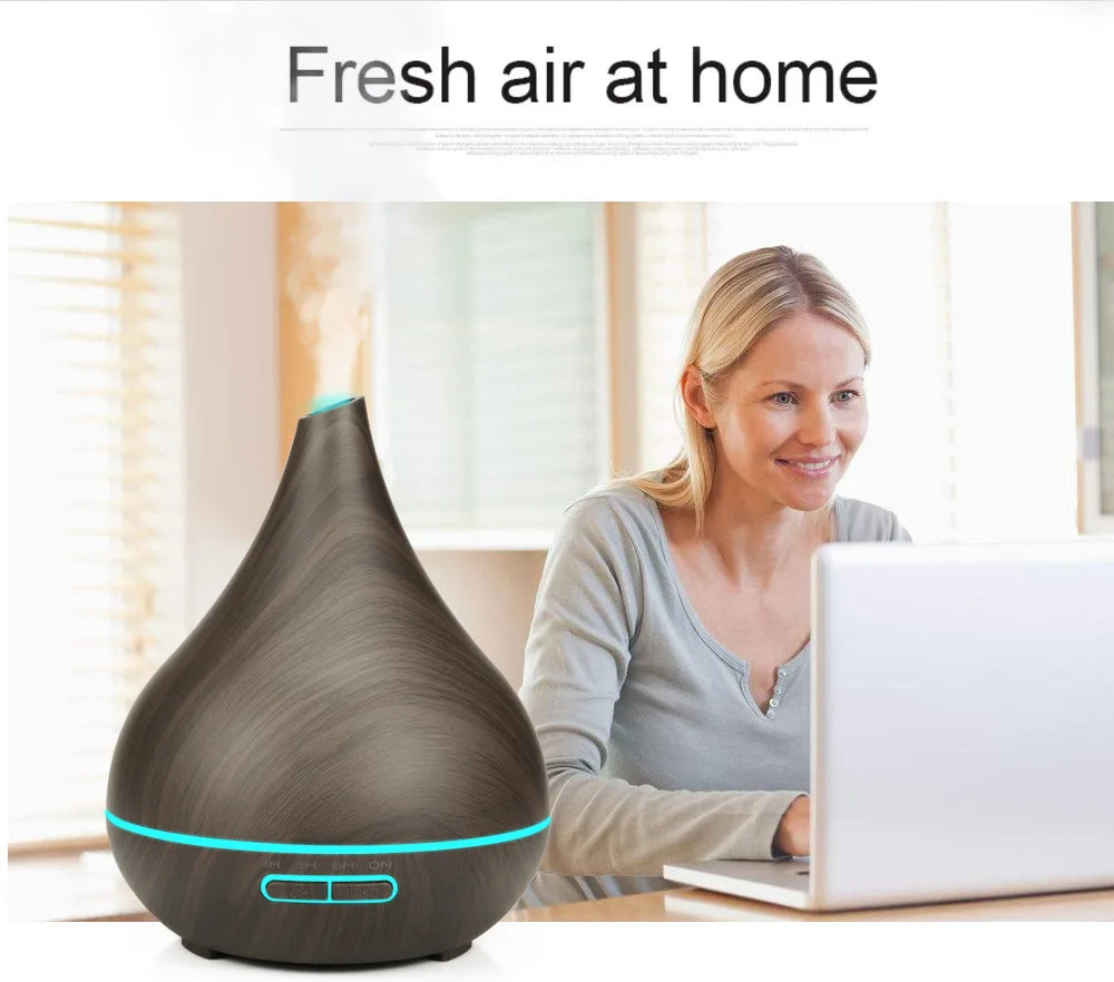 Dark wood-grain teardrop diffuser with a glowing blue light band, emitting mist on a desk near a woman using a laptop.