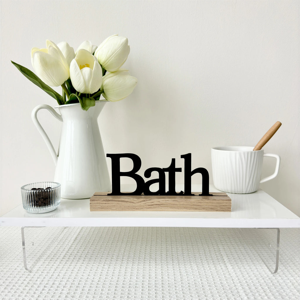 Black 'Bath' sign on a wooden base, displayed on a white shelf with white tulips in a jug and a textured mug.