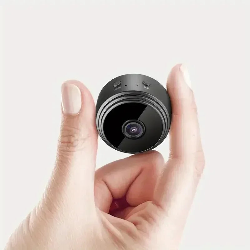 A small, black, circular plastic security camera held between thumb and finger, showing a lens and two buttons.