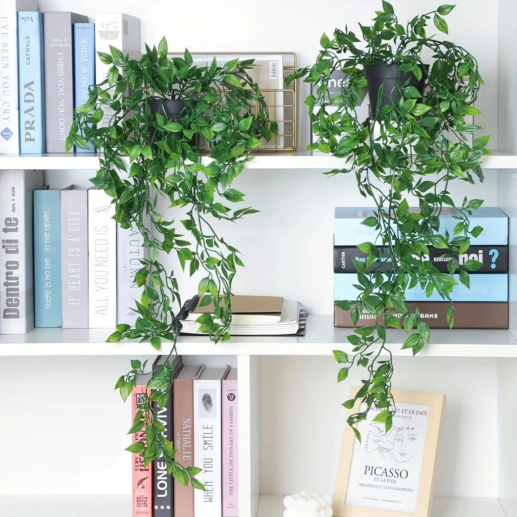 Two artificial hanging ivy plants in black pots, displayed on white shelves filled with decorative books and art.