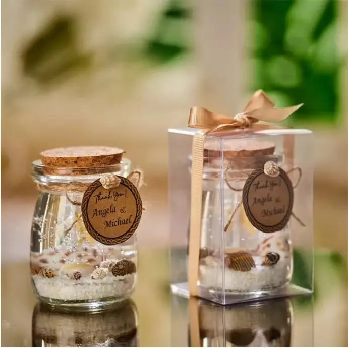 Two glass jars with cork lids, filled with sand and seashells, featuring personalized "Thank You" tags and ribbon bows.
