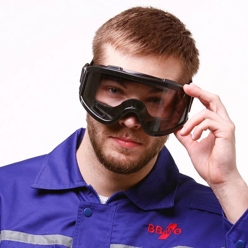 Black anti-impact safety goggles with clear wrap-around lenses worn by a man in a blue work shirt.