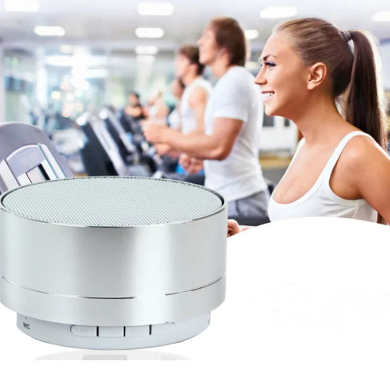 Compact silver aluminium Bluetooth speaker with a mesh top, sitting in front of a woman exercising in a gym.