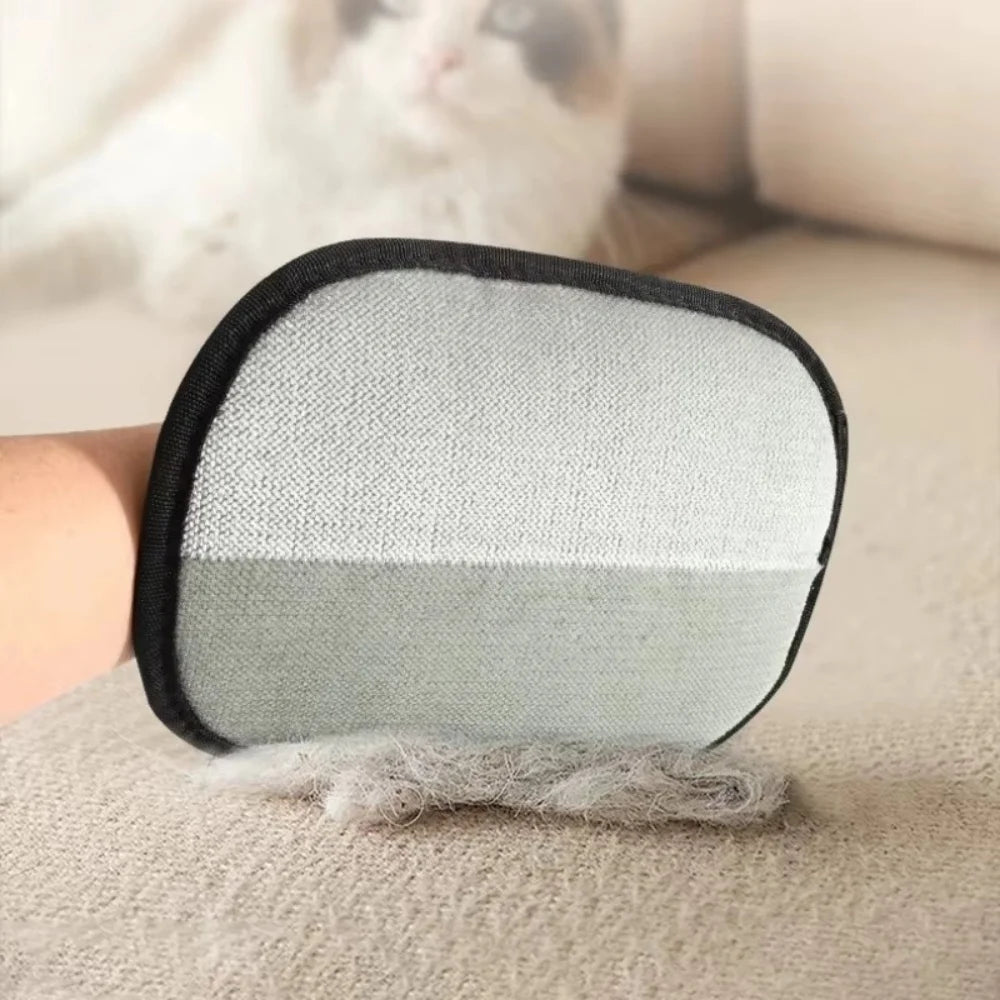 A hand uses a grey, rectangular fabric pet hair remover mitt to clean a sofa, with a fluffy cat visible behind.