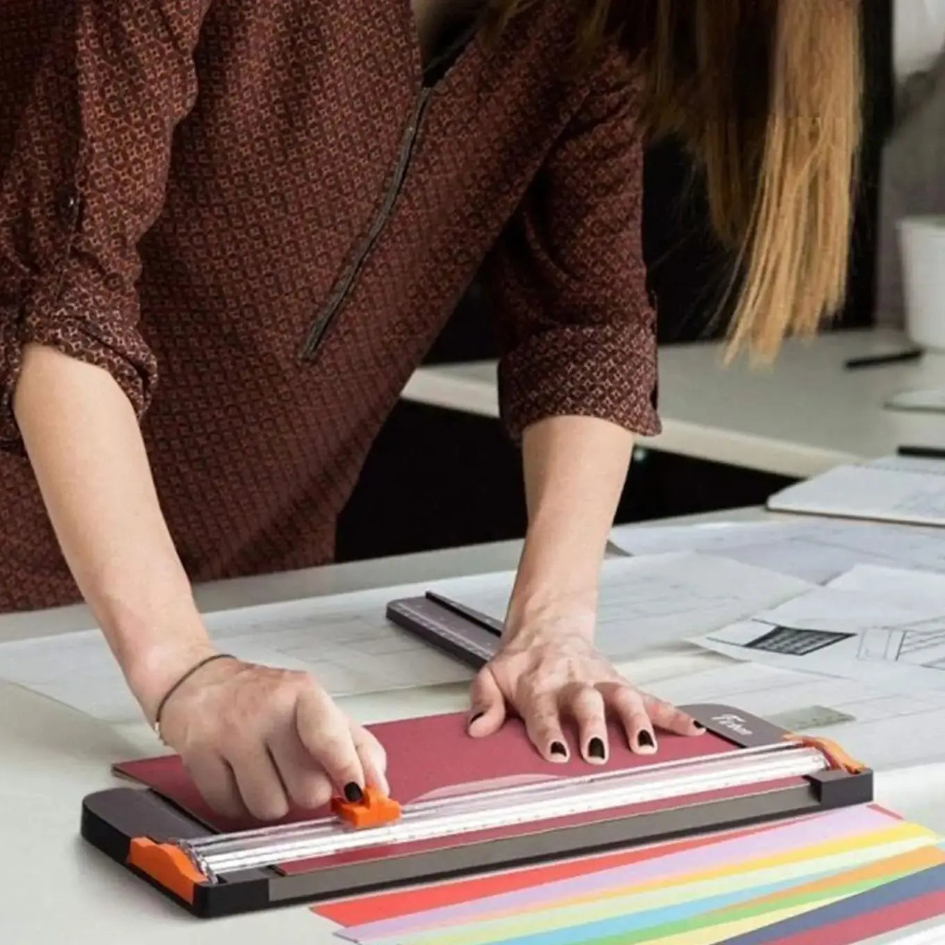 Person using a black paper trimmer with an orange sliding blade to cut a red sheet on a desk with multi-coloured paper.