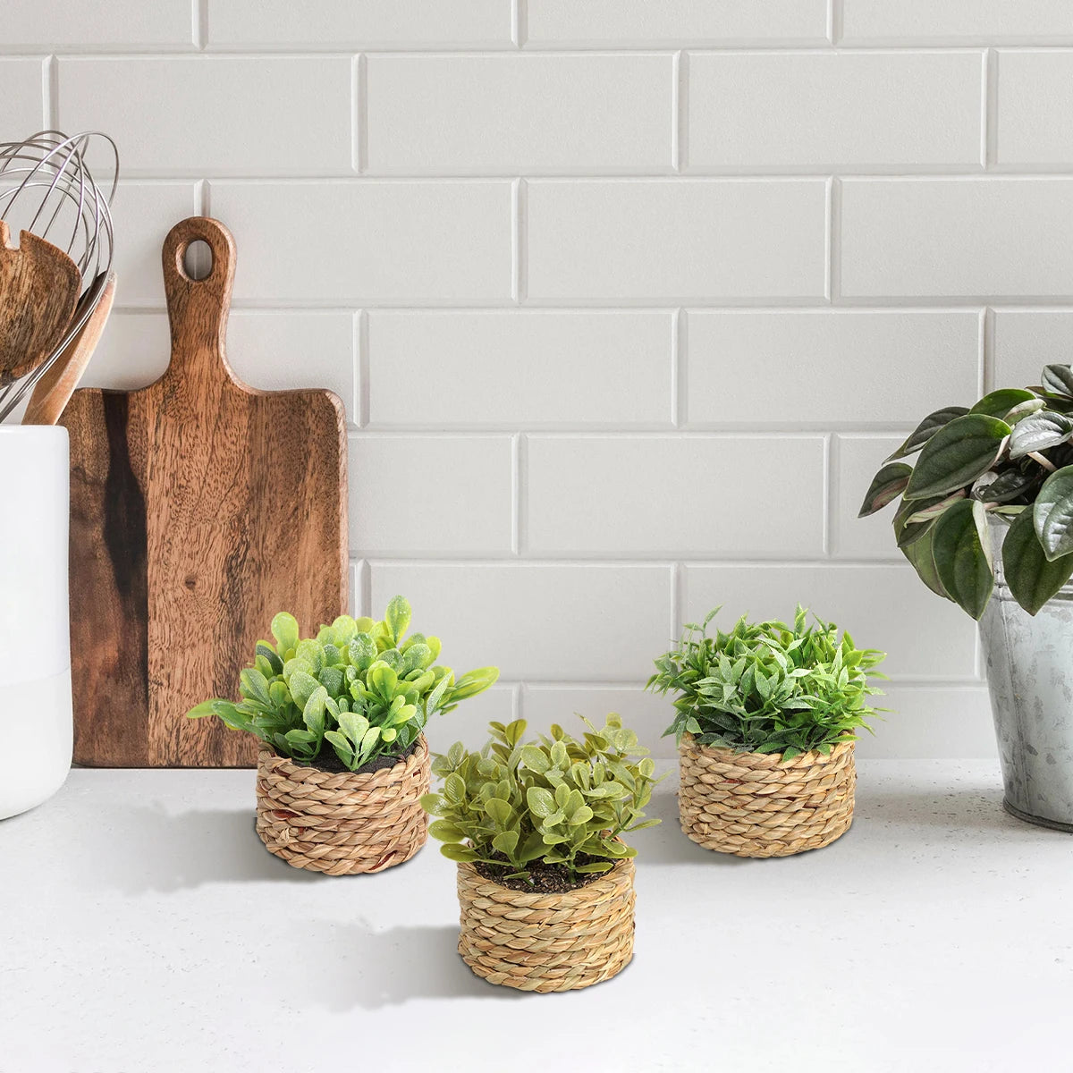 Three small artificial green plants in woven grass pots sit on a kitchen counter against a white subway tile backsplash.