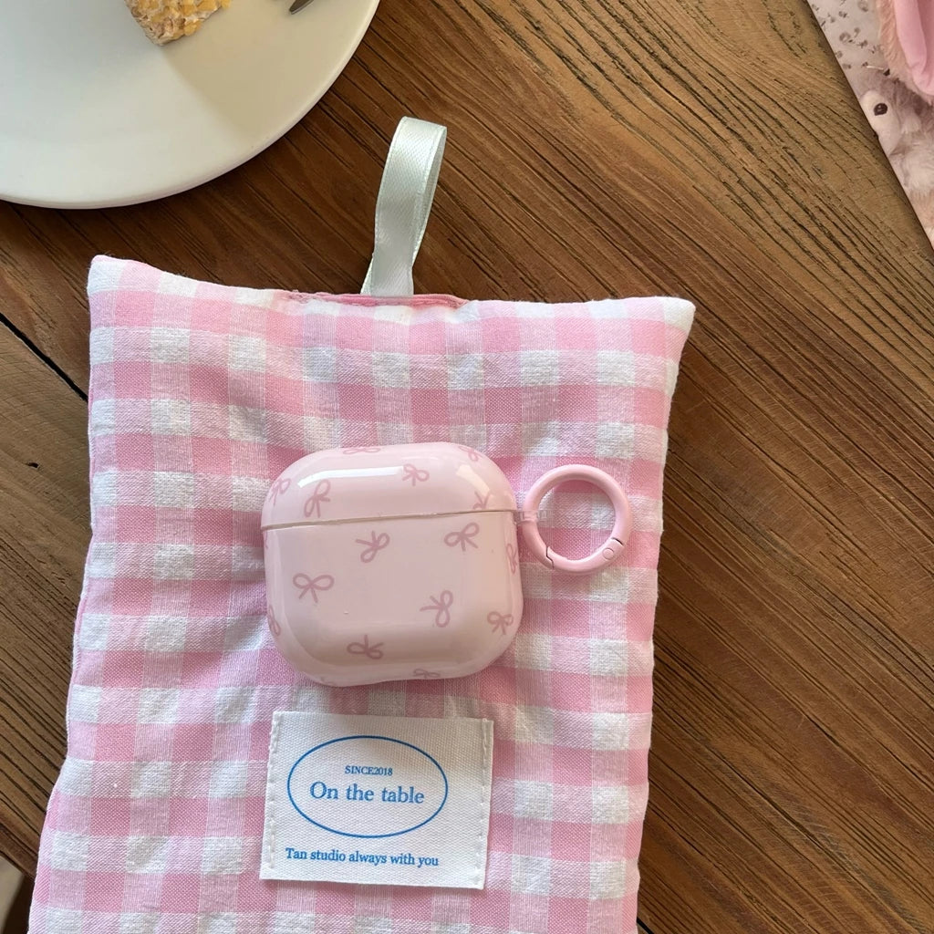 Glossy pale pink AirPods case with white bow pattern and ring clip, resting on a pink gingham fabric pouch.