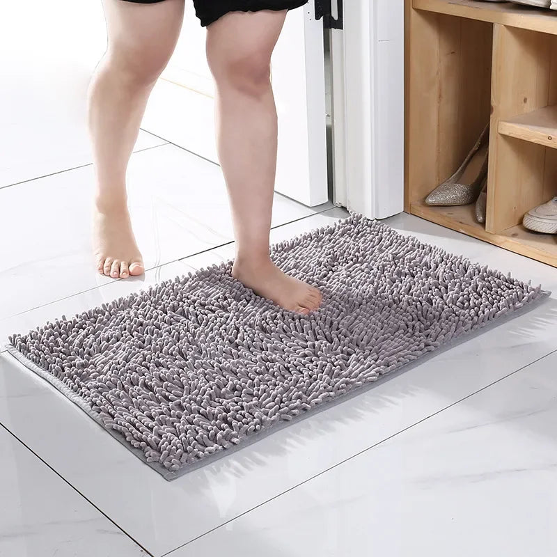 Person standing on a rectangular, grey, shaggy-textured absorbent doormat for muddy paws on a tiled floor.