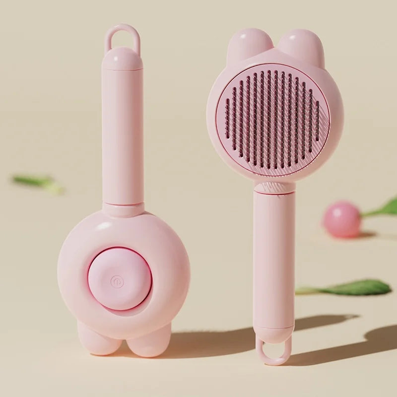 Two baby pink, cat-ear-shaped pet grooming brushes with a self-cleaning button, set against a cream background.