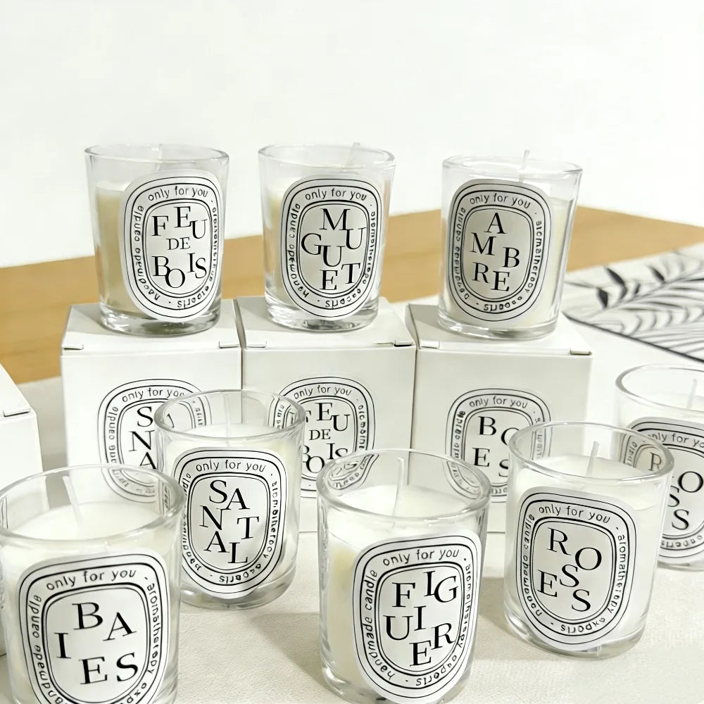 Assorted white scented candles in clear glass votives with minimalist oval labels and matching white gift boxes.
