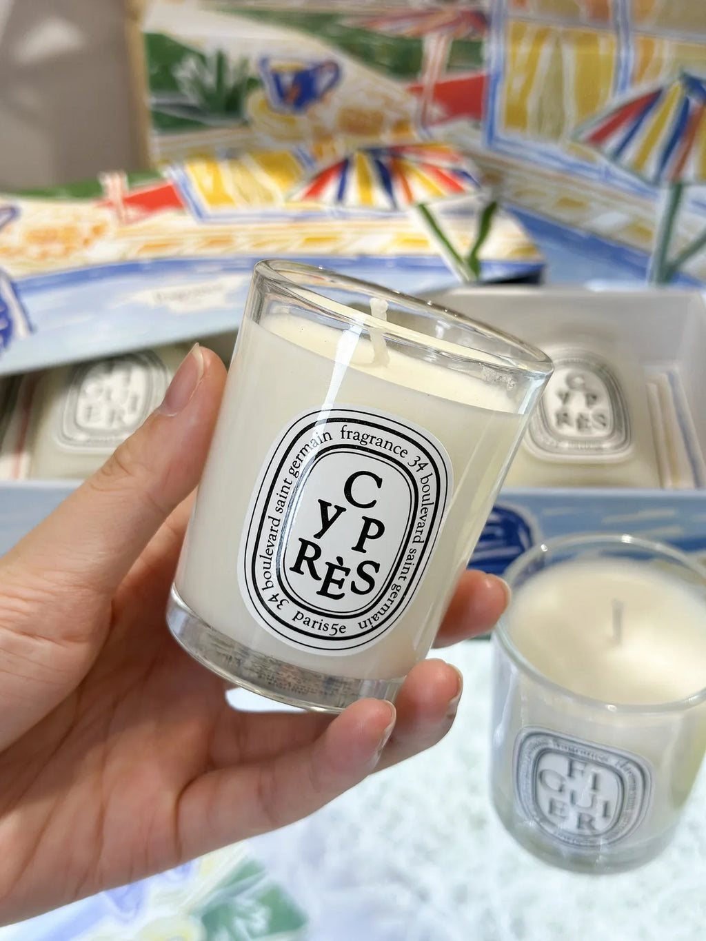 Hand holding a white scented candle in a clear glass jar with a decorative oval label, set in a gift box.