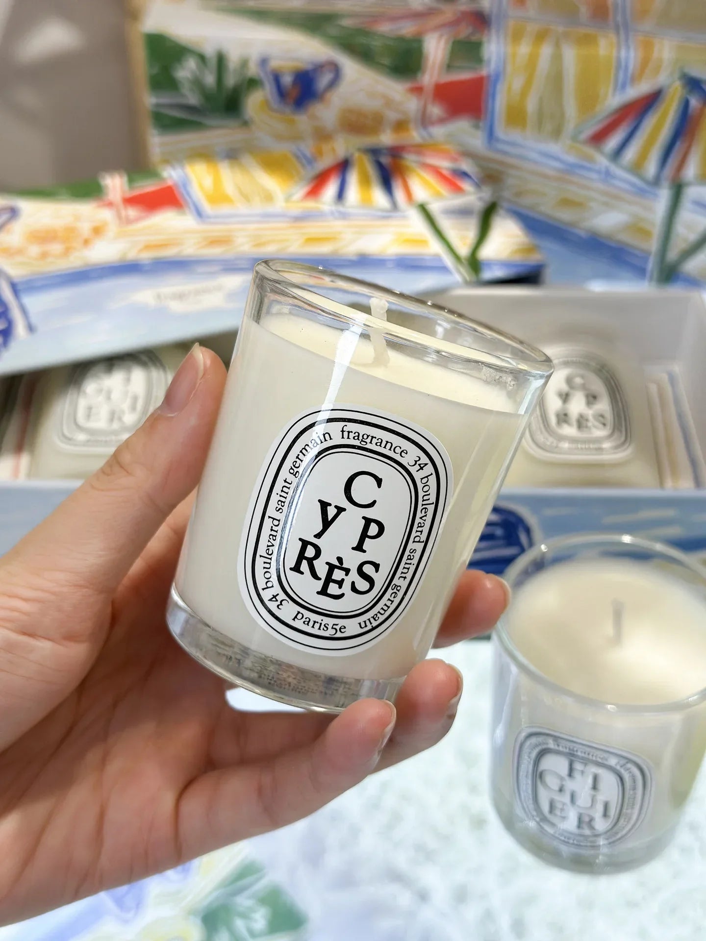 Hand holding a white scented candle in a clear glass jar with a decorative oval label, set in a gift box.