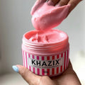 A hand holding an open 100g pot of thick, pink intensive body cream with a striped label.