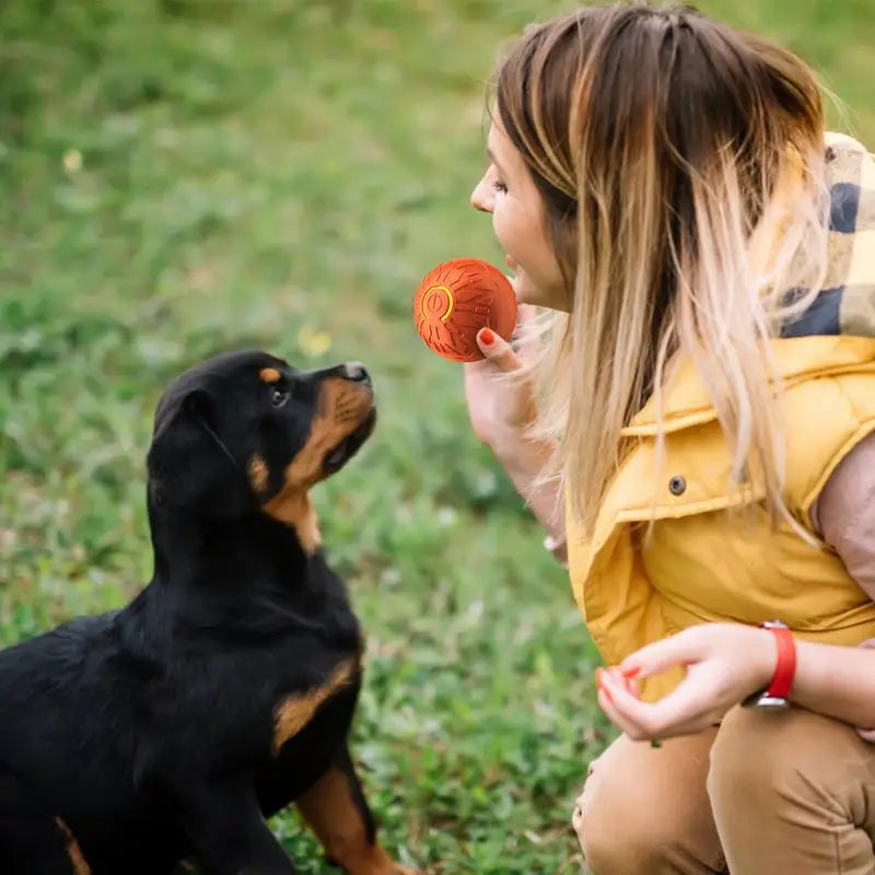 Woman holding an orange, textured automatic interactive dog toy ball while playing with a puppy on the grass.