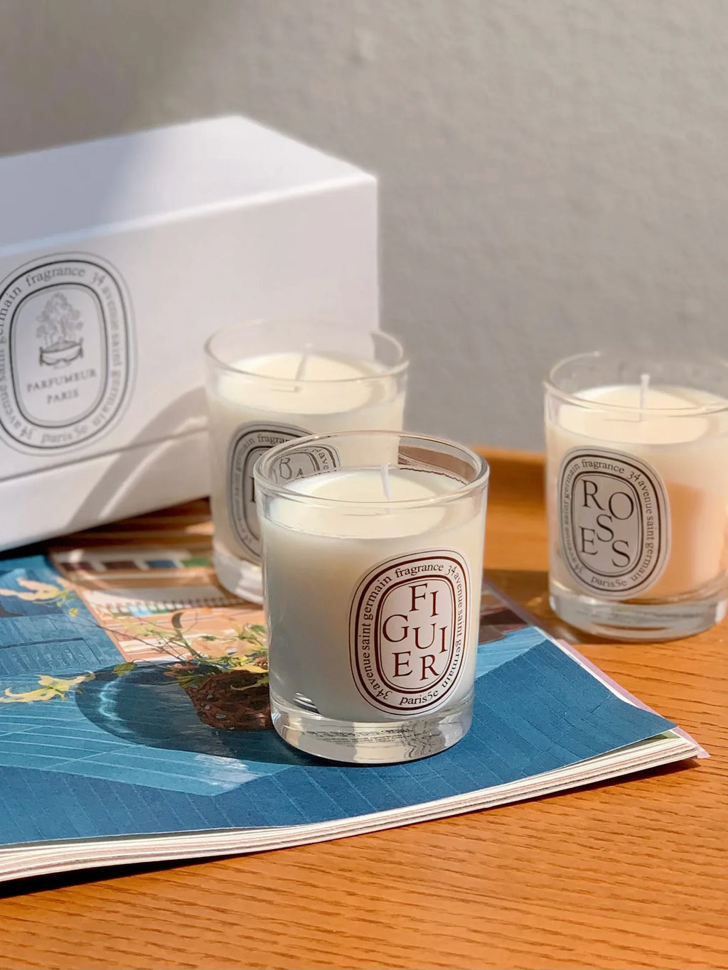 Three scented candles in glass jars with white wax and oval labels, arranged on a magazine on a wooden surface.