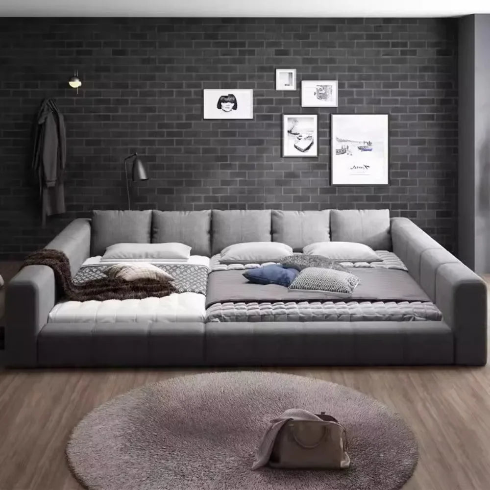 Modern grey padded double bed with high, wraparound cushioned sides, set against a dark brick wall.