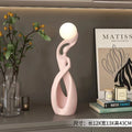 Abstract pale pink resin sculpture featuring a smooth, swirling silhouette topped with a glowing white spherical light.