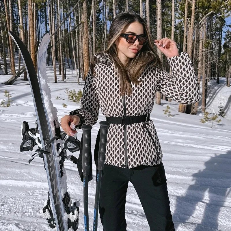 Woman in a brown and white diamond-patterned quilted ski jacket with a black waist belt, posing with skis and poles.