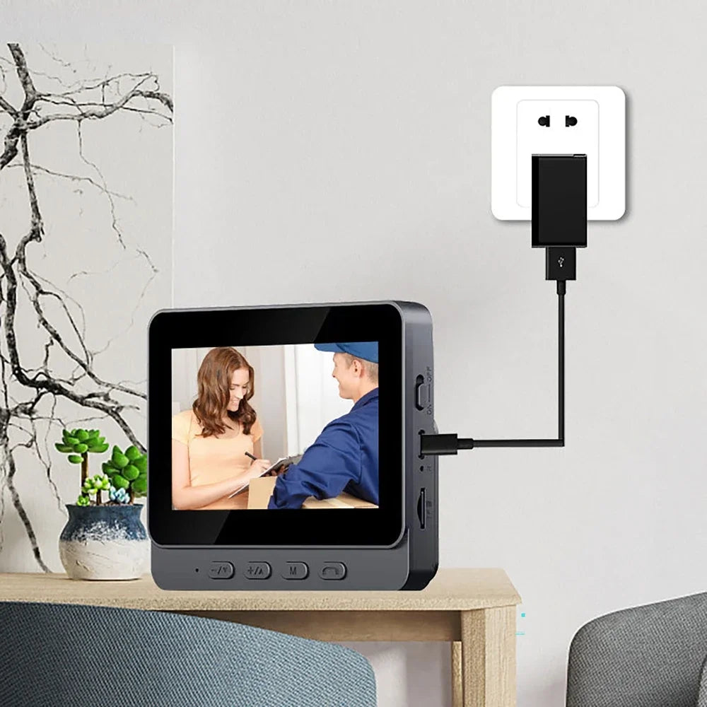 A sleek, dark grey smart video doorbell with a 4.3-inch HD screen, plugged into a wall outlet on a wooden table.