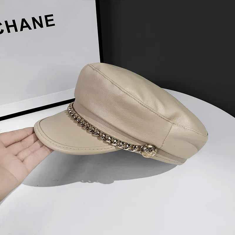 Beige faux leather baker boy cap featuring a decorative gold chain and button detail along the peak.