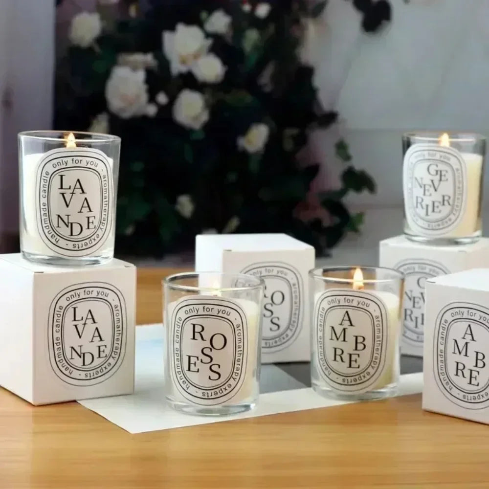 A group of lit scented candles in clear glass jars with white, oval minimalist labels on a wooden table.