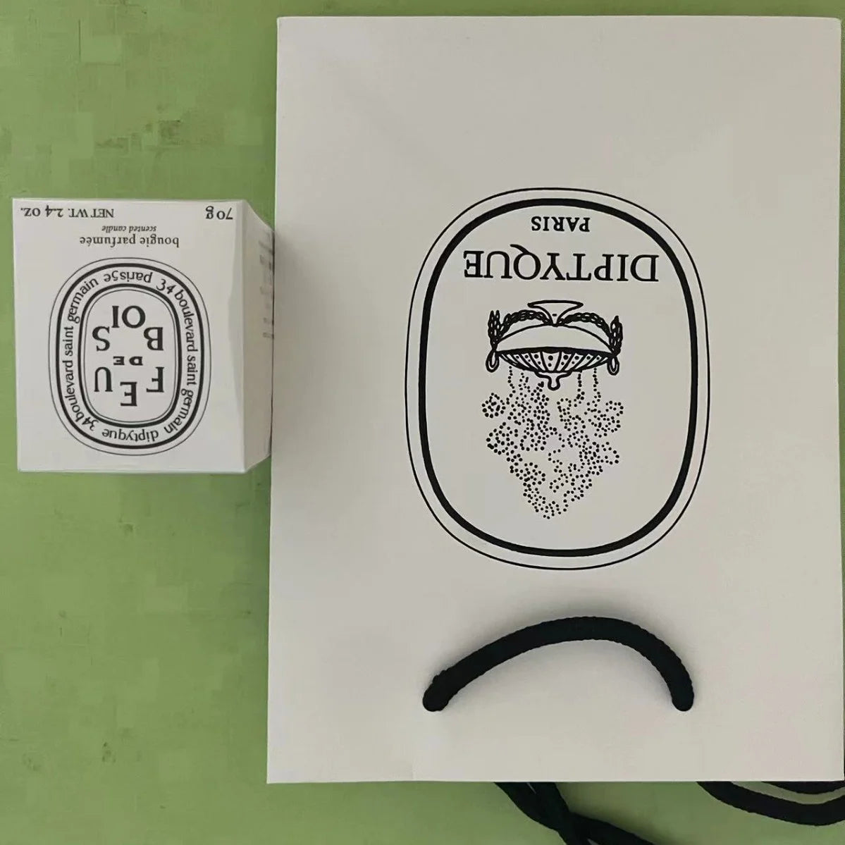 A white scented candle box and matching branded white gift bag with black handles and ornate graphic design.