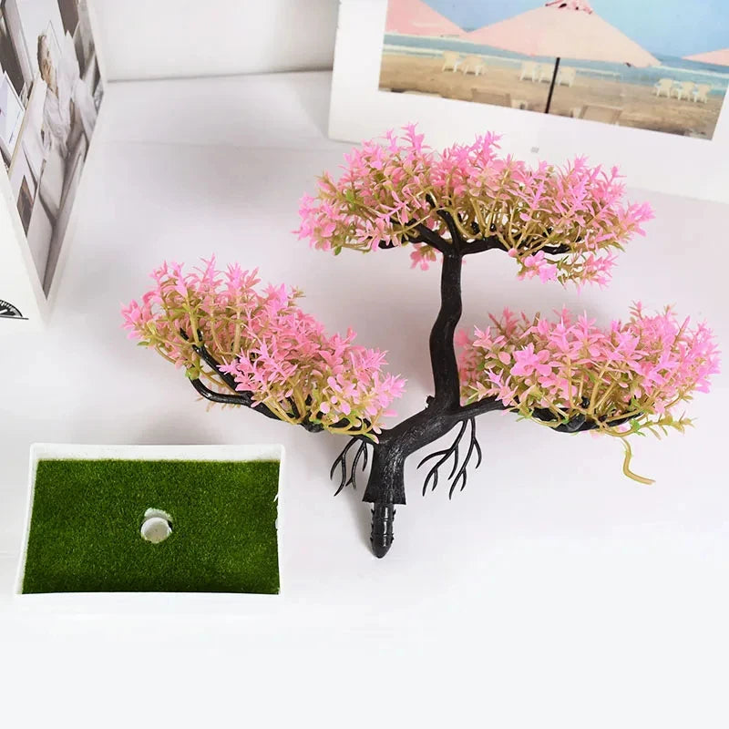Artificial bonsai tree with pink leaves and black branches, next to a small, empty white rectangular pot with moss.
