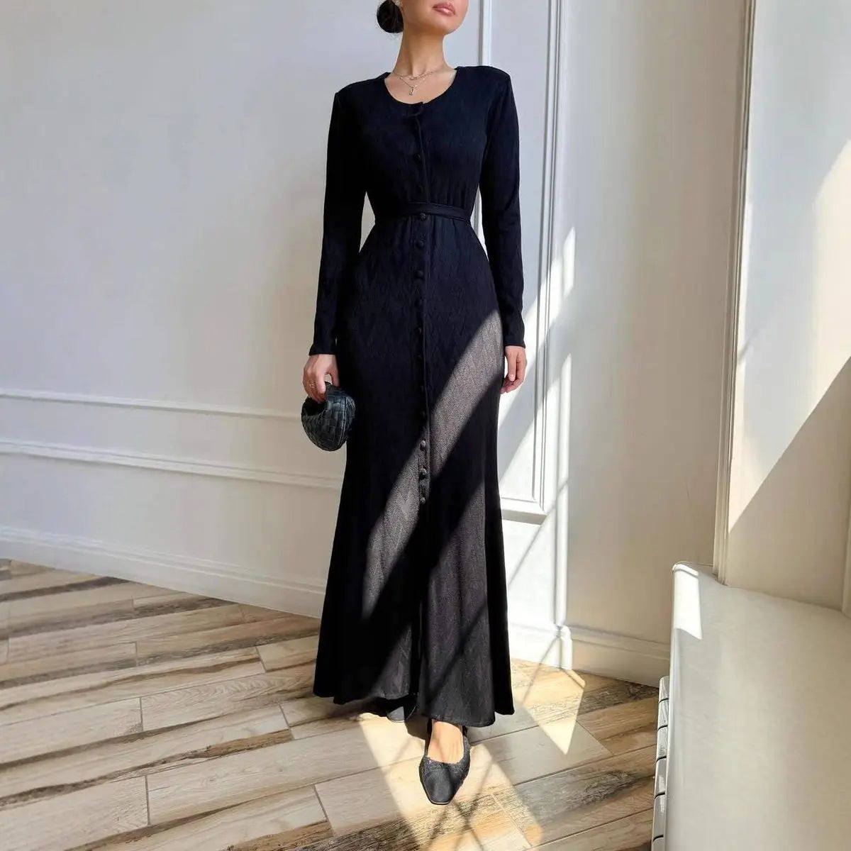 Elegant black long-sleeved maxi dress with a button-down front, cinched waist, and textured fabric finish.