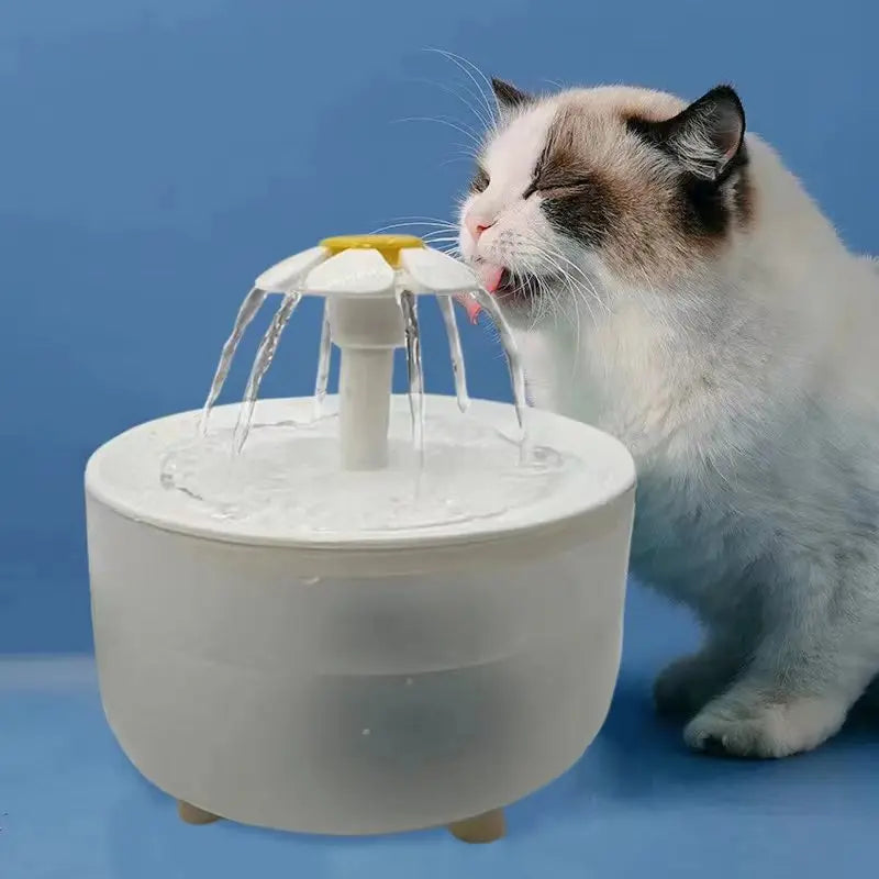 A white, round pet water fountain with a daisy-shaped flower top, featuring a cat drinking from the cascading water.