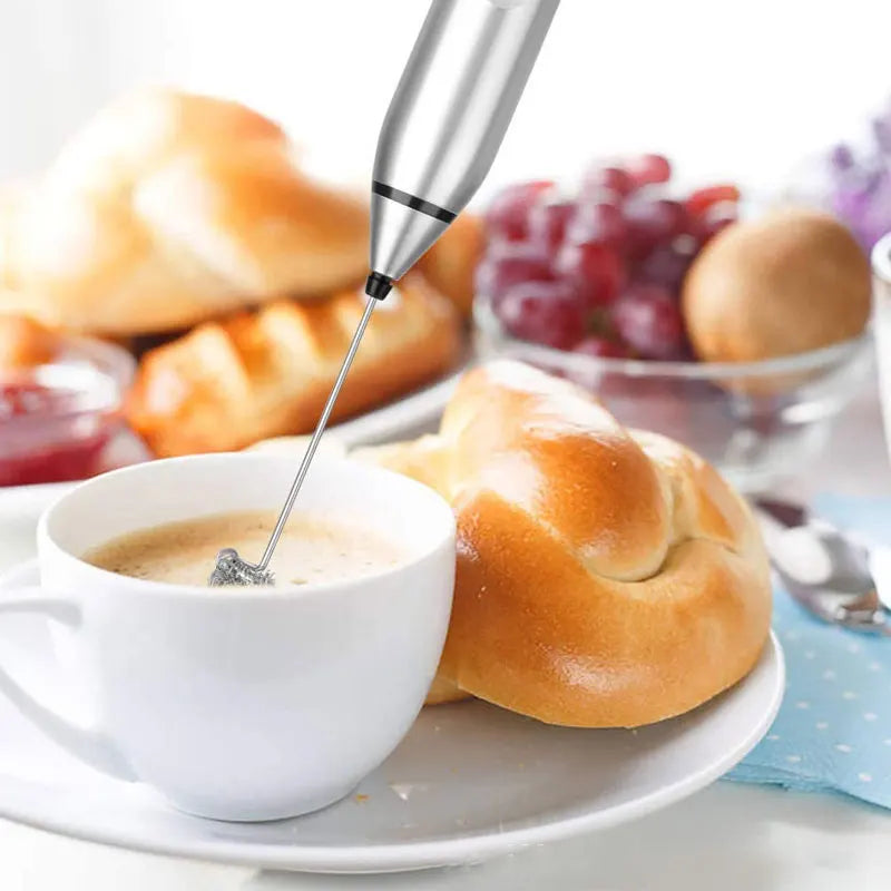 A stainless steel handheld electric milk frother mixing coffee in a white cup, on a table with breakfast pastries.