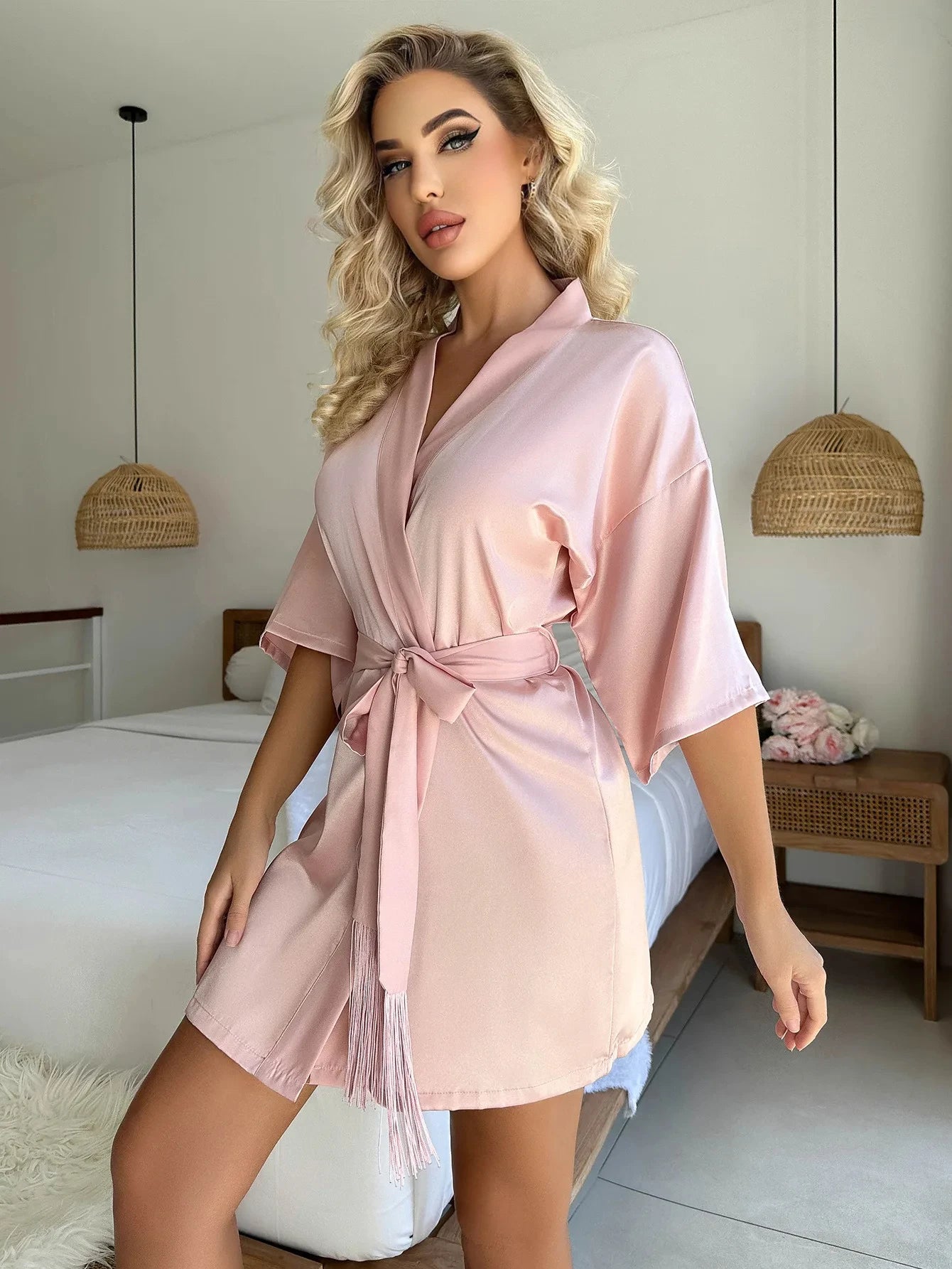 Woman wearing a short, blush pink satin robe with wide sleeves, a tie waist, and elegant tassel detailing.