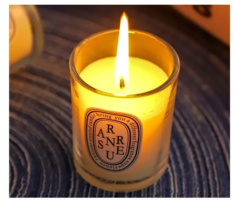 A glowing scented candle in a clear glass votive jar with a decorative oval label on a dark textured background.