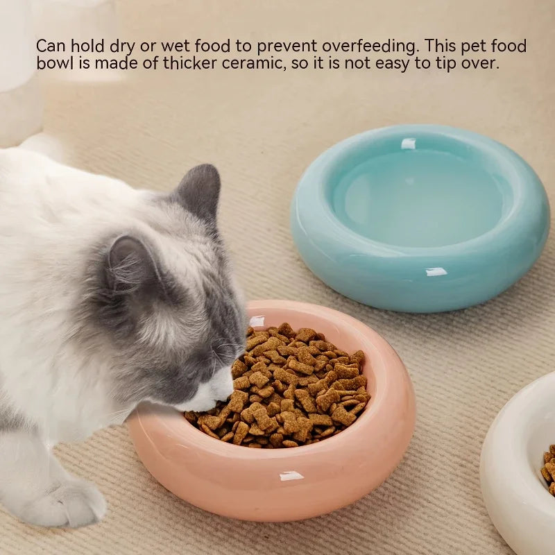 A fluffy cat eats from a rounded, pale pink ceramic bowl, with blue and white bowls nearby on a neutral surface.