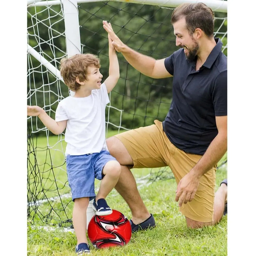A young boy and his father high-fiving by a goal post, with a red and black patterned football on the grass.