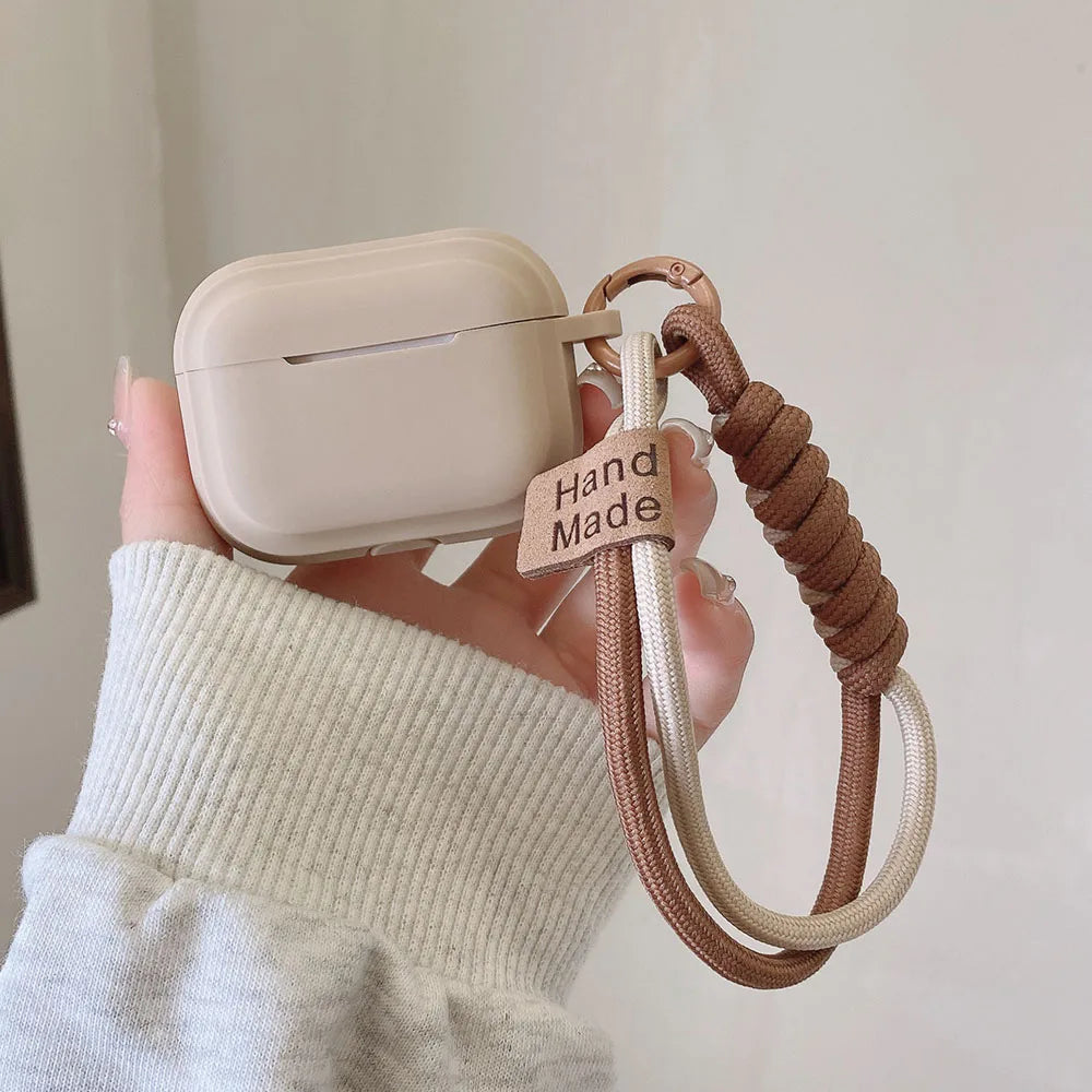 Beige AirPods Pro case with a handmade braided cord wrist strap, held in a hand against a plain background.