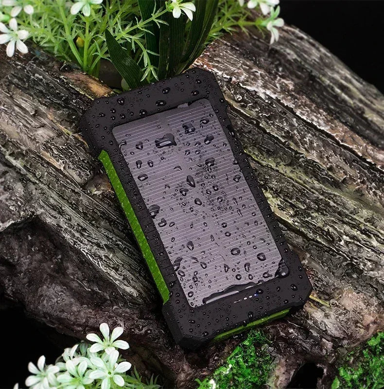 Rugged black and green portable solar power bank with water droplets, resting on mossy bark with white flowers.