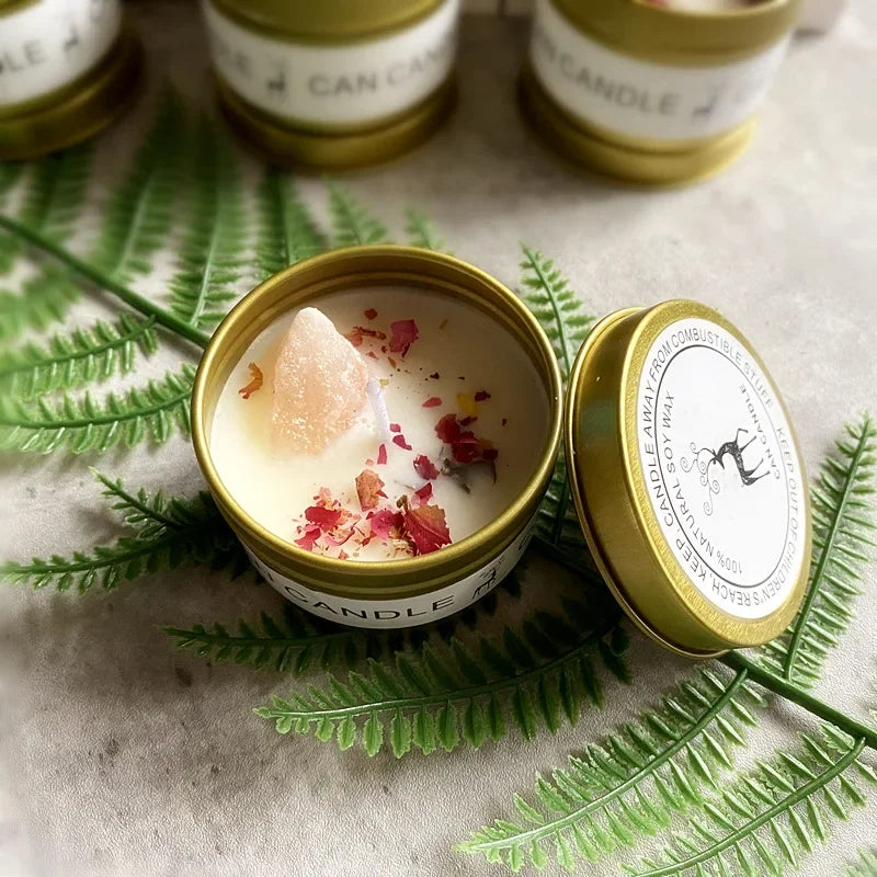 Scented soy candle in a gold tin, topped with a raw crystal and dried flower petals, resting on a fern leaf.