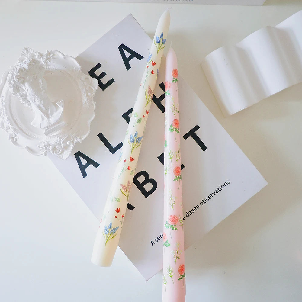 Two tall tapered dinner candles, one cream with blue flowers and one pink with orange roses, on a white background.